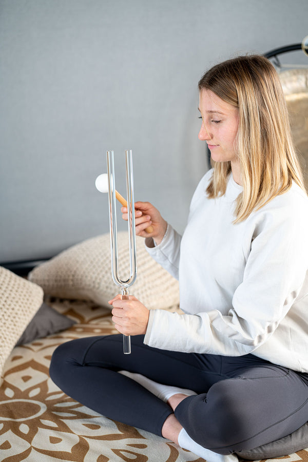 A woman is sitting on a bed with Meinl Crystal Tuning Fork 174.61 HZ / F3, 17.7" in her hand, experimenting with the sound produced when striking them with a hammer.