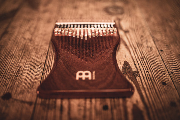 A melodic instrument, the wooden ukulele, is laying on top of a Meinl 17 notes Kalimba, Sapele, on a wooden table.