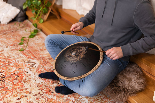 A man sitting on the floor playing a Meinl 16" Tongue Drum, D Kurd, Lasered floral design, Black, made of high-quality stainless steel.