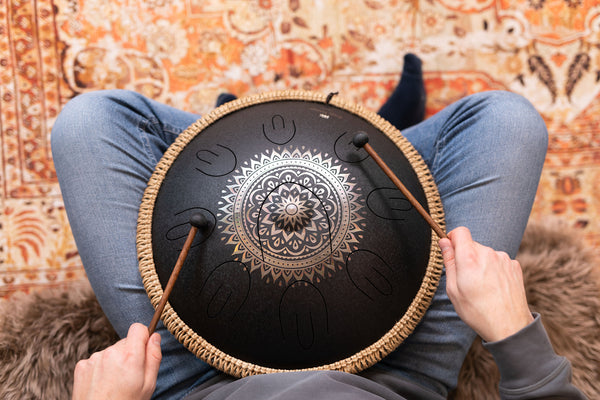 A person sitting on a rug holding a Meinl 16" Tongue Drum, D Kurd, Lasered floral design, Black, creating meditative sound with this high-quality stainless steel instrument.