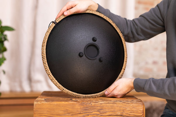 A man is gently striking the Meinl 16" Tongue Drum, D Kurd, Lasered floral design, Black, creating meditative sounds. He skillfully balances the black disc on top of a wooden table.