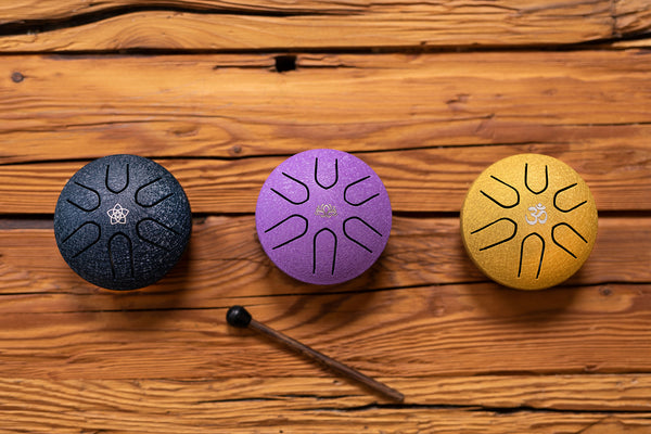 Three different colored chimes, including the Meinl Pocket Steel Tongue Drum (3" Tongue Drum, A Major, OM, Gold), sitting on top of a wooden table.